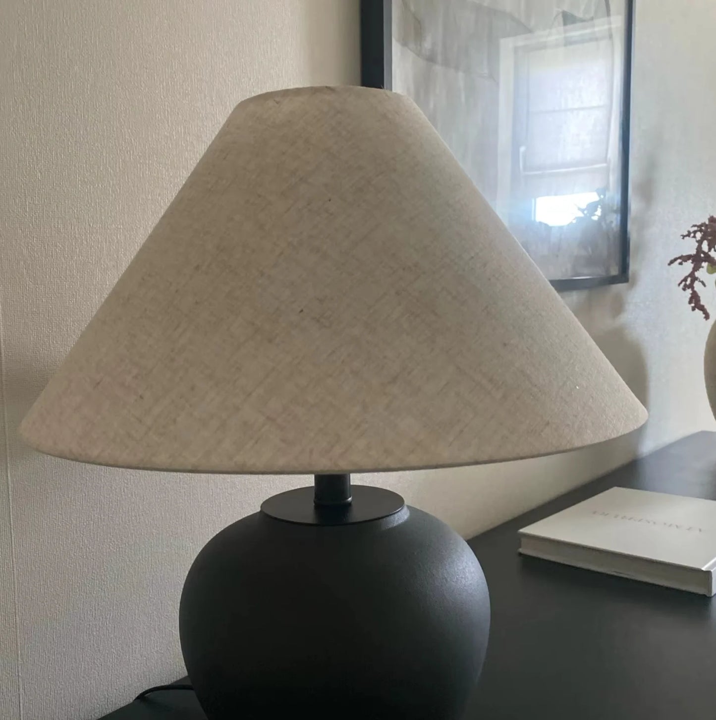 Table lamp with beige shade and black base on a surface, with a book in the background.