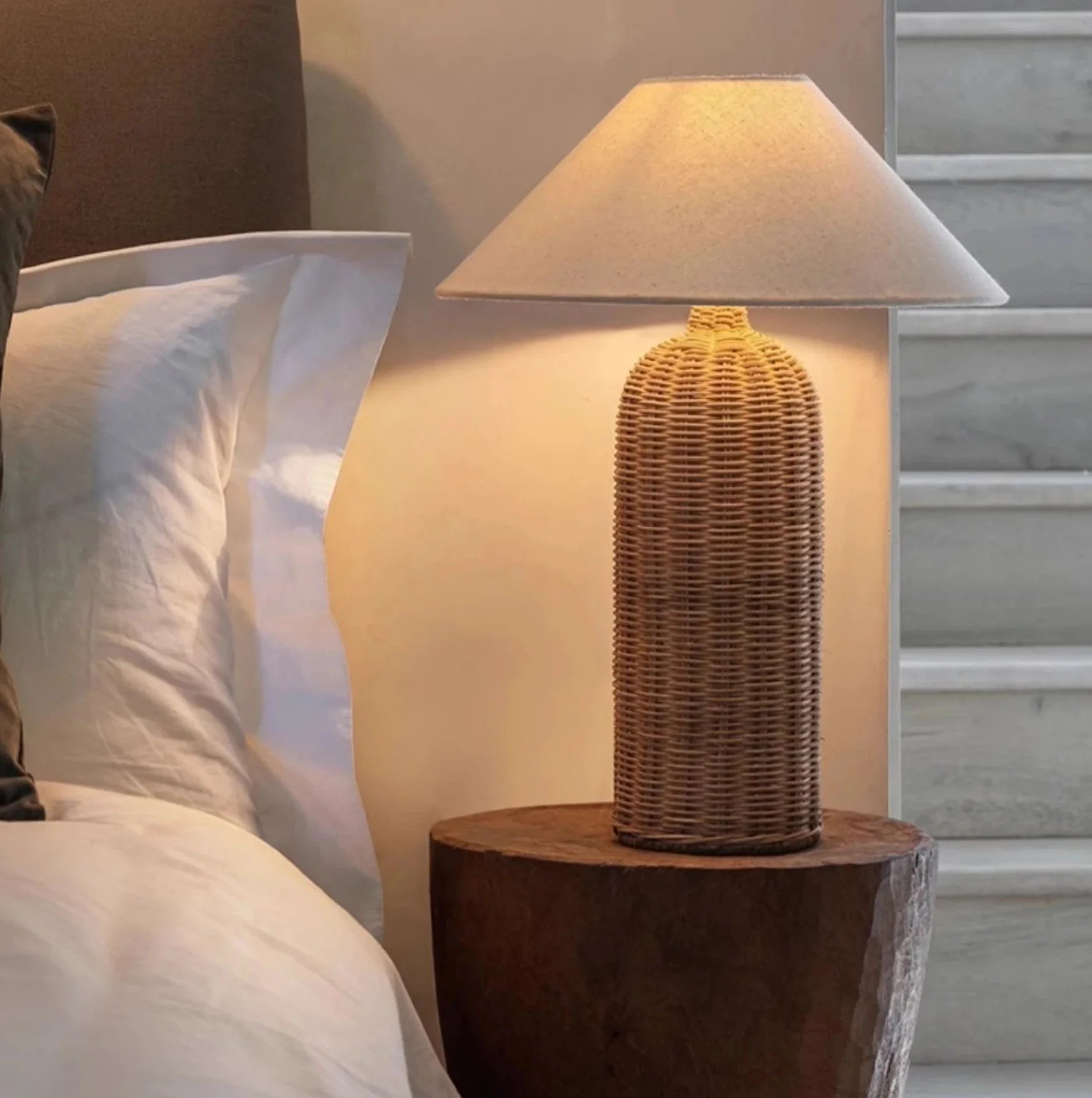 Wicker table lamp on a wooden stand next to a bed with pillows