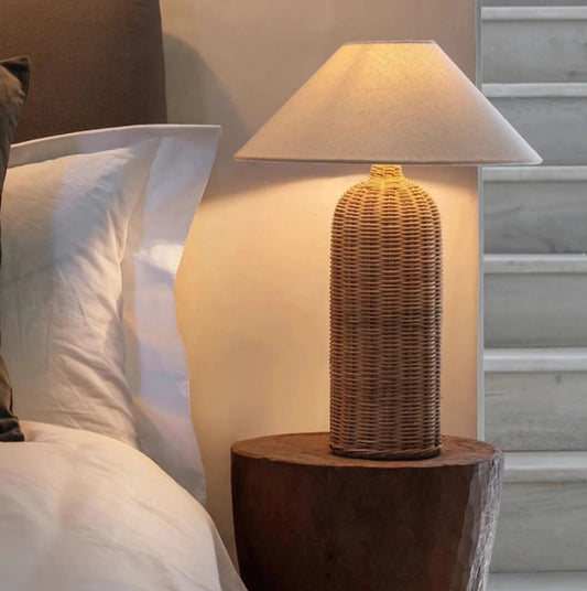 Wicker table lamp on a wooden stand next to a bed with pillows