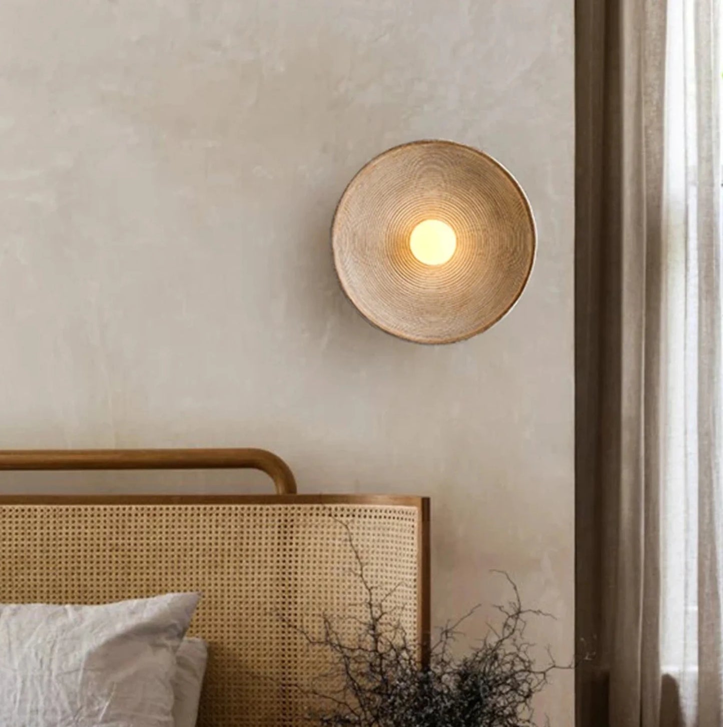 Ribbed round wall light fixture on a neutral wall above a bed with wicker headboard.