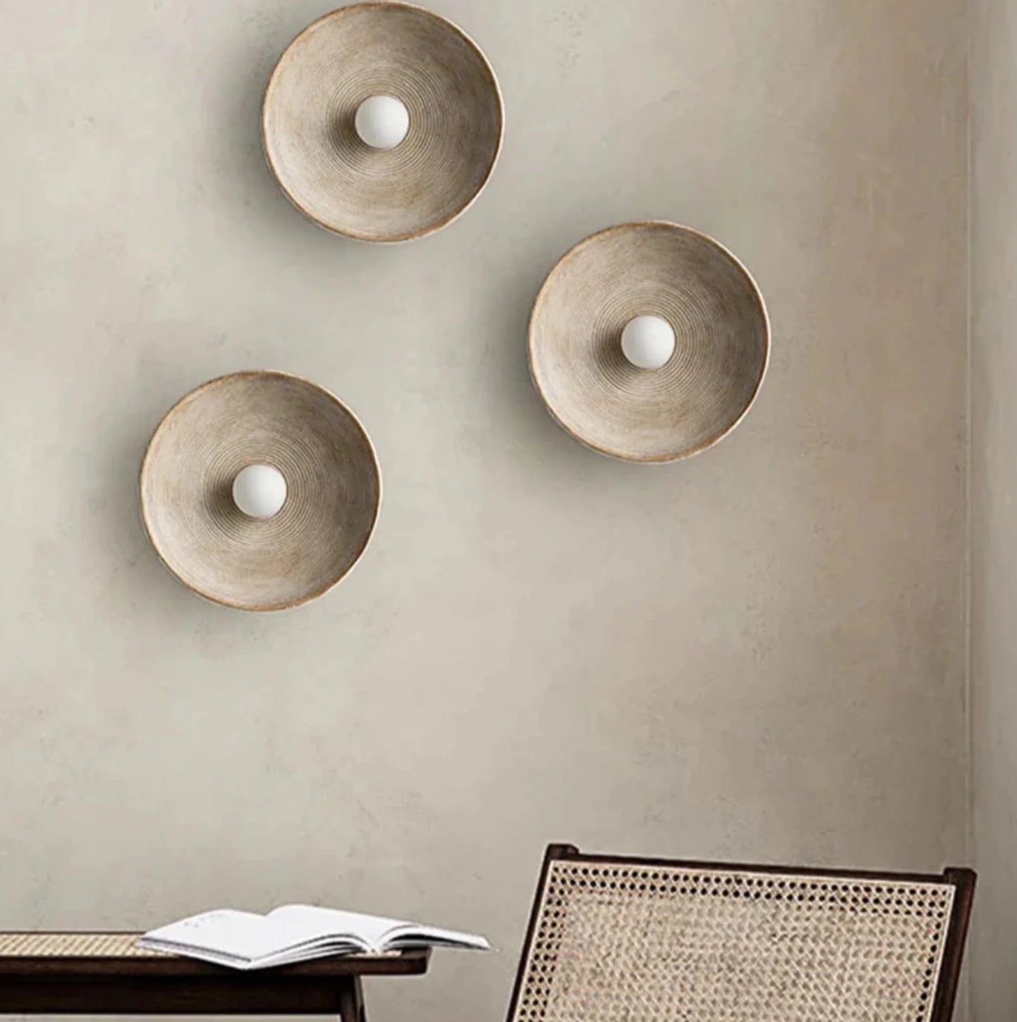 Three wall-mounted round l wall light fixtures on a beige wall with a chair and open book in the foreground.