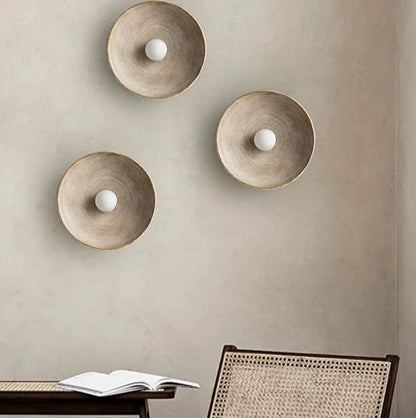 Three wall-mounted round l wall light fixtures on a beige wall with a chair and open book in the foreground.