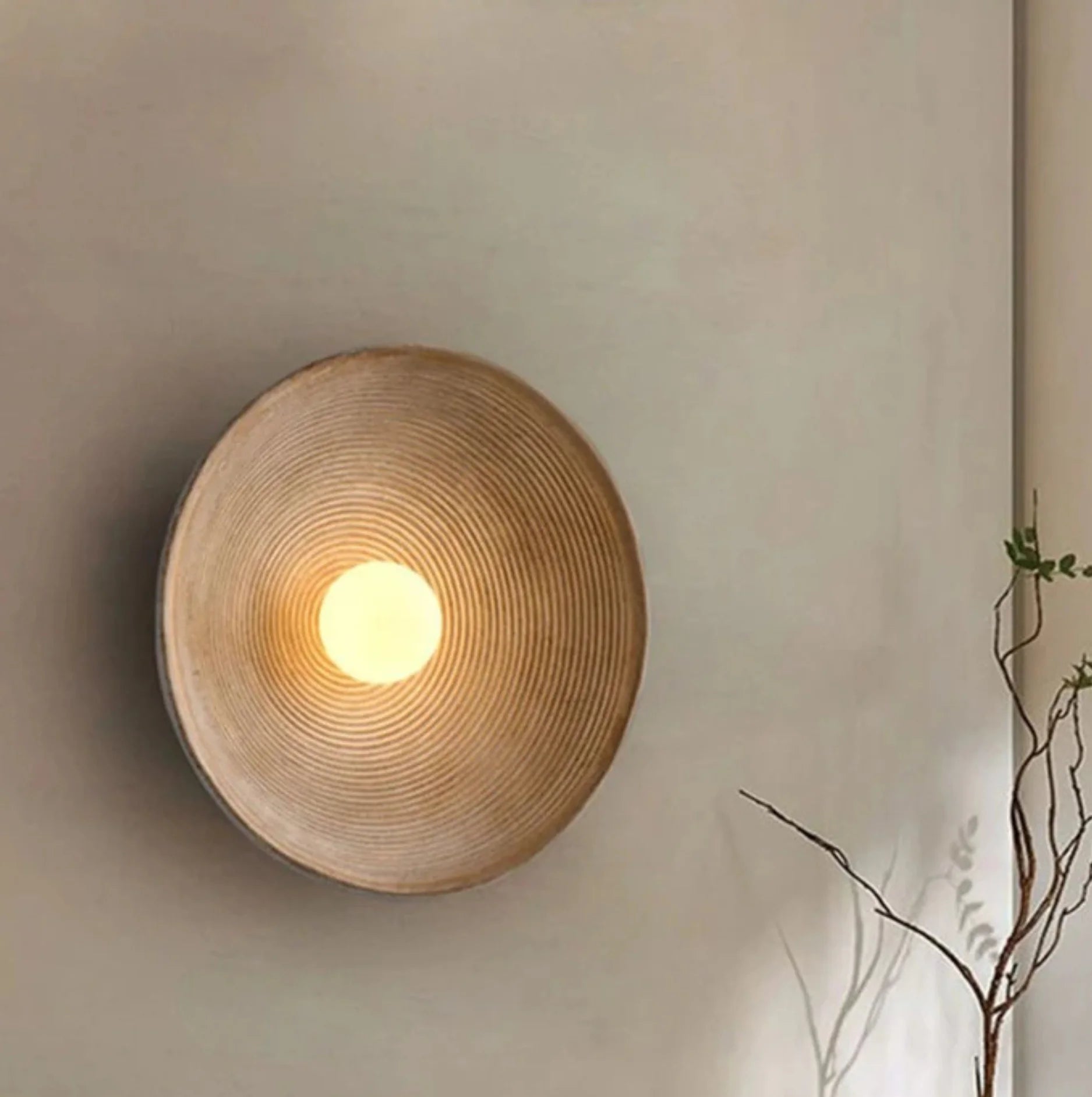 Round woven wall light fixture on a neutral wall with a plant branch in the corner.